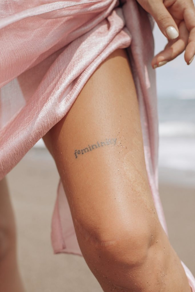 A photo of a woman's leg with a silk pink dress and a tattoo on the leg saying "femininity."
