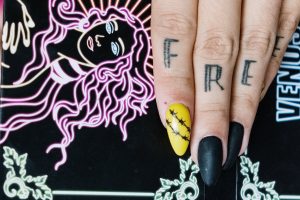 A womans hand with black and yellow nails and a tattoo on her fingers saying "Free"
