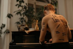 A guy playing a piano with naked back full of text tattoos