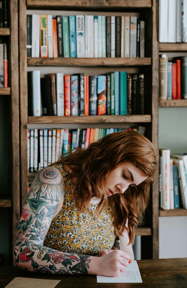 Woman writing on white paper with Sunflower shoulder tattoo and bookshelf in the background