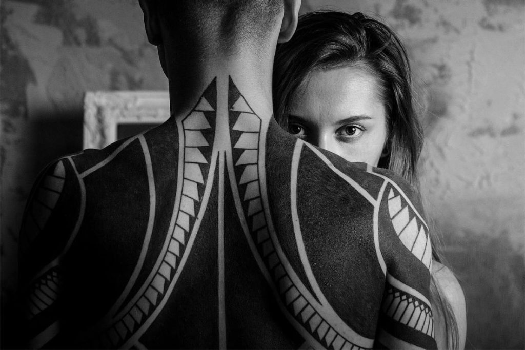 A woman hiding behind man with a big cover up tattoo on his back
